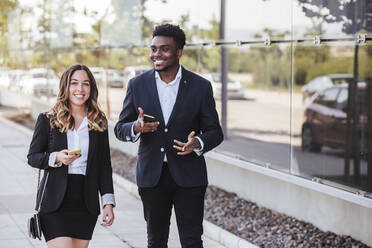 Smiling business professionals holding smart phones while walking on sidewalk in city - LJF01650