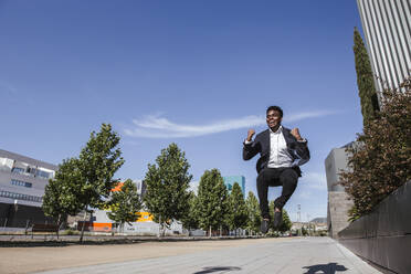 Cheerful male professional jumping on footpath against blue sky in city during sunny day - LJF01656