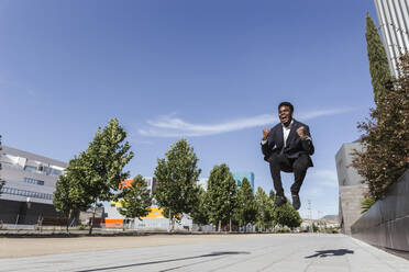 Cheerful businessman jumping on footpath against blue sky in city during sunny day - LJF01657
