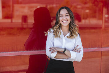 Lächelnde Geschäftsfrau mit verschränkten Armen vor einer roten Wand in der Stadt - LJF01665