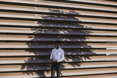 Smiling businessman standing against modern built structure with shadow of tree - LJF01674