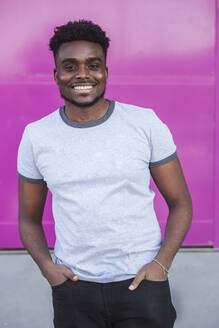 Smiling young man with hands in pockets standing against pink wall - LJF01675