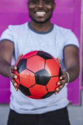 Close-up of young man holding soccer ball while standing against pink color - LJF01676