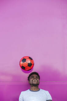 Fröhlicher junger Mann jongliert mit Fußball gegen eine rosa Wand in der Stadt - LJF01680