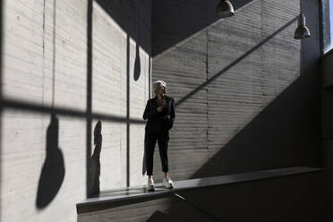 Geschäftsfrau in elegantem Anzug steht auf Stützmauer mit Sonnenlicht und Schatten im Hintergrund - TCEF00811