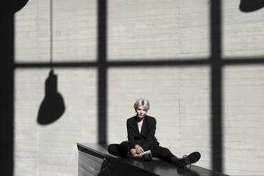 Female professional wearing black suit relaxing against sunlight and shadow in background at office - TCEF00814