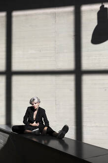 Stilvolle Geschäftsfrau in elegantem Anzug entspannt sich auf einer Stützmauer mit Sonnenlicht und Schatten im Hintergrund - TCEF00815