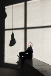 Junge Frau in elegantem Anzug sitzt mit Sonnenlicht und Schatten im Hintergrund im Büro - TCEF00820