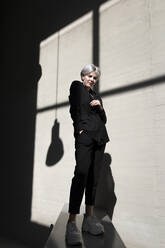 Stilvolle Frau in elegantem Anzug steht auf Stützmauer mit Sonnenlicht und Schatten im Hintergrund im Büro - TCEF00822