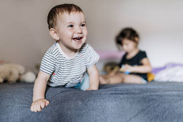 Porträt eines glücklichen kleinen Mädchens, das auf dem Bett krabbelt, während ihre Schwester im Hintergrund sitzt - GEMF03896