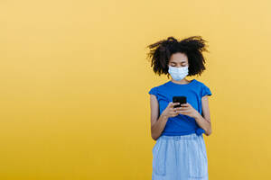 Young woman wearing protective mask, using smartphone in front of yellow wall - TCEF00840