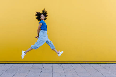 Pretty woman jumping for joy in front of yellow wall - TCEF00841