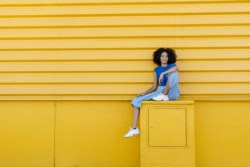 Pretty woman sitting on platform in front of yellow wall, smiling - TCEF00847