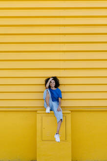 Hübsche Frau sitzt auf einem Podest vor einer gelben Wand und lächelt - TCEF00848