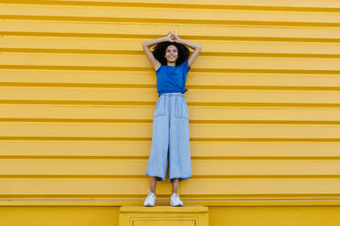 Smiling woman standing on platform in front of yellow wall - TCEF00849