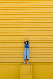 Smiling woman reaching up at yellow wall, standing on platform - TCEF00850