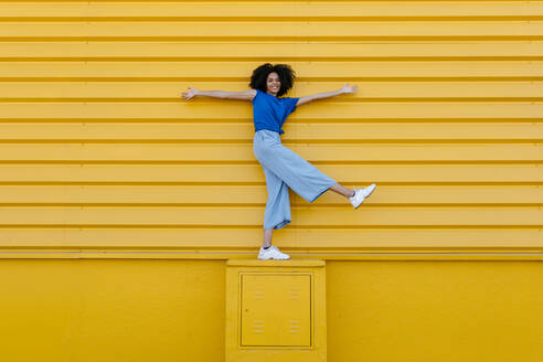 Happy young woman balancing on platform in front of yellow wall - TCEF00851