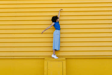 Happy young woman balancing on platform in front of yellow wall - TCEF00852