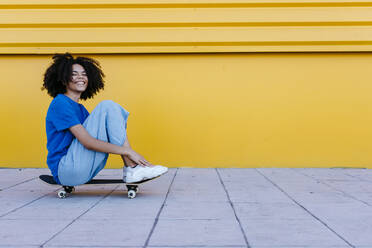 Lächelnde junge Frau sitzt auf einem Skateboard vor einer gelben Wand - TCEF00858