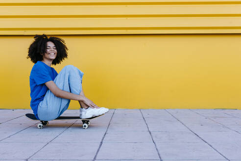 Smiling young woman sitting on skateboard in front of yellow wall - TCEF00858