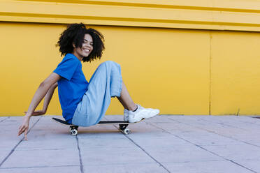 Lächelnde junge Frau sitzt auf einem Skateboard vor einer gelben Wand - TCEF00859