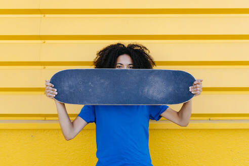 Young woman holding skateboard in front of face, looking at camera - TCEF00864