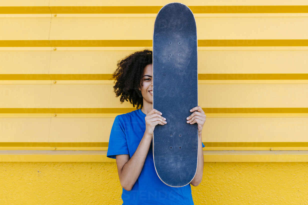 Young woman holding skateboard in front of face, looking at camera