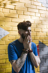 Young man with tattoo standing in front of yellow brick wall, covering mouth with his hands - EYAF01191