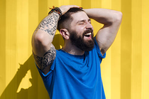 Lachender junger Mann mit Tattoo vor einer gelben Wand - EYAF01198