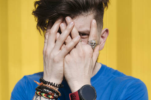 Young man standing in front of yellow wall, hiding face behind his hands - EYAF01199