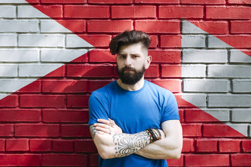 Young man with tattoo standing in front of red brick wall with arms crossed - EYAF01202