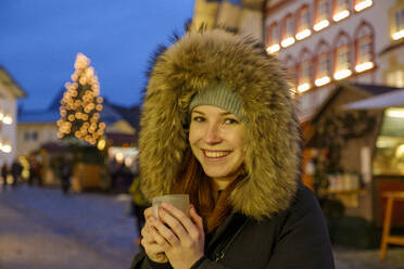 Happy woman wearing fur hood while holding coffee cup at Christmas market - LBF03132