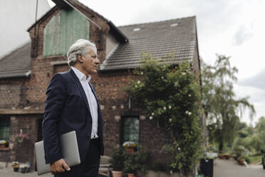 Porträt eines älteren Geschäftsmannes mit Laptop vor einem Bauernhaus - GUSF04019