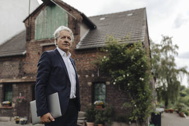 Portrait of a senior businessman holding laptop in front of a farmhouse - GUSF04021