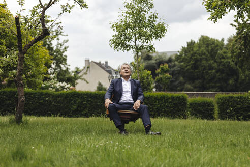 Älterer Geschäftsmann sitzt auf einem Stuhl in einem ländlichen Garten und schaut nach oben - GUSF04044