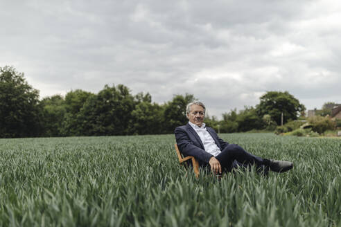 Älterer Geschäftsmann sitzt auf einem Stuhl in einem Feld auf dem Lande - GUSF04068
