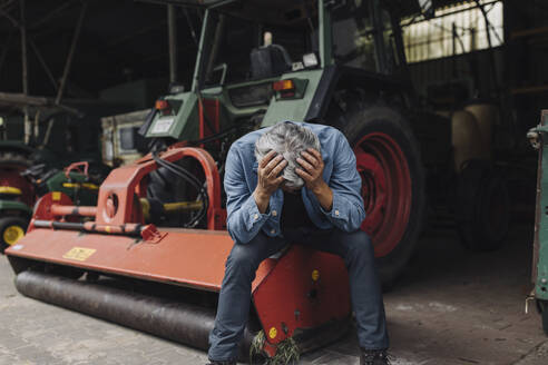 Verzweifelter älterer Mann auf einem Bauernhof, der auf einem Traktor in einer Scheune sitzt - GUSF04102