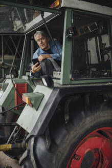 Älterer Mann mit Smartphone auf einem Traktor auf einem Bauernhof - GUSF04116