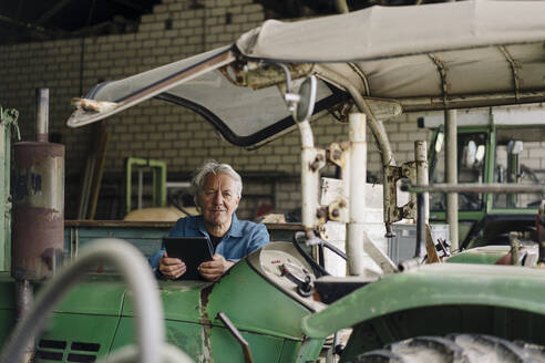 Senior man holding tablet on a farm with tractor in barn - GUSF04119