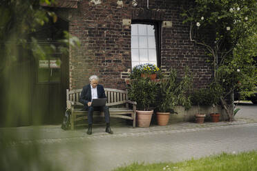 Senior businessman sitting at a farmhouse using laptop - GUSF04134