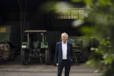 Porträt eines älteren Geschäftsmannes mit Laptop auf einem Bauernhof mit Traktor in einer Scheune, lizenzfreies Stockfoto