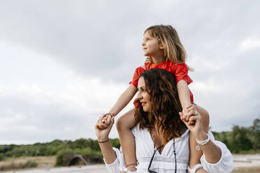 Thoughtful woman carrying daughter on shoulders against cloudy sky - EGAF00375