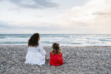 Mother and daughter relaxing on shore at beach against cloudy sky - EGAF00383