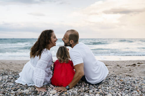 Paar küsst sich gerade, während es mit seiner Tochter am Strand sitzt - EGAF00385