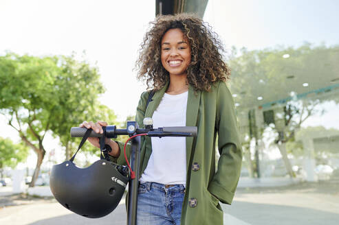 Glückliche junge Frau mit Elektro-Scooter auf der Straße stehend - KIJF03123