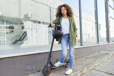 Confident young woman standing with electric push scooter on sidewalk - KIJF03126