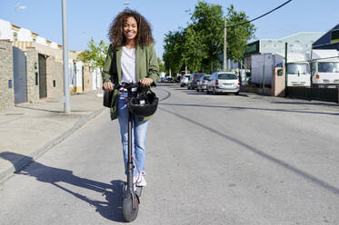 Glückliche Frau fährt elektrischen Roller auf der Straße an einem sonnigen Tag - KIJF03132