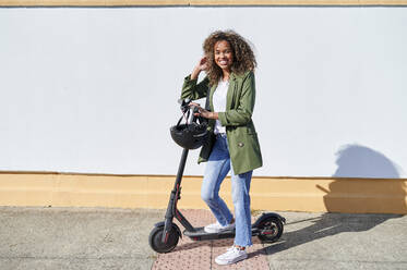 Glückliche Frau steht mit elektrischem Tretroller auf dem Bürgersteig an einem sonnigen Tag - KIJF03133