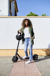 Confident woman standing with electric push scooter on sidewalk during sunny day - KIJF03135