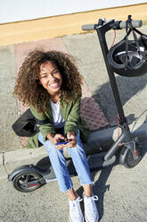 Happy young woman holding smart phone while sitting on sidewalk with electric push scooter - KIJF03137